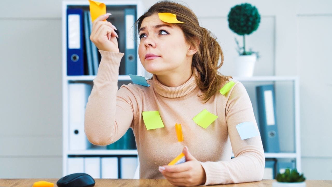 A woman sitting at a desk with sticky notes on her head, organizing her tasks for the day.