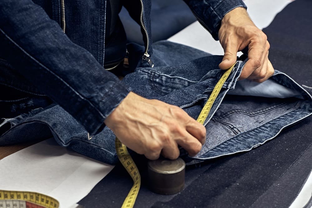 Close-up of a person measuring denim fabric with a yellow measuring tape, wearing a denim jacket, with pattern materials on the table.